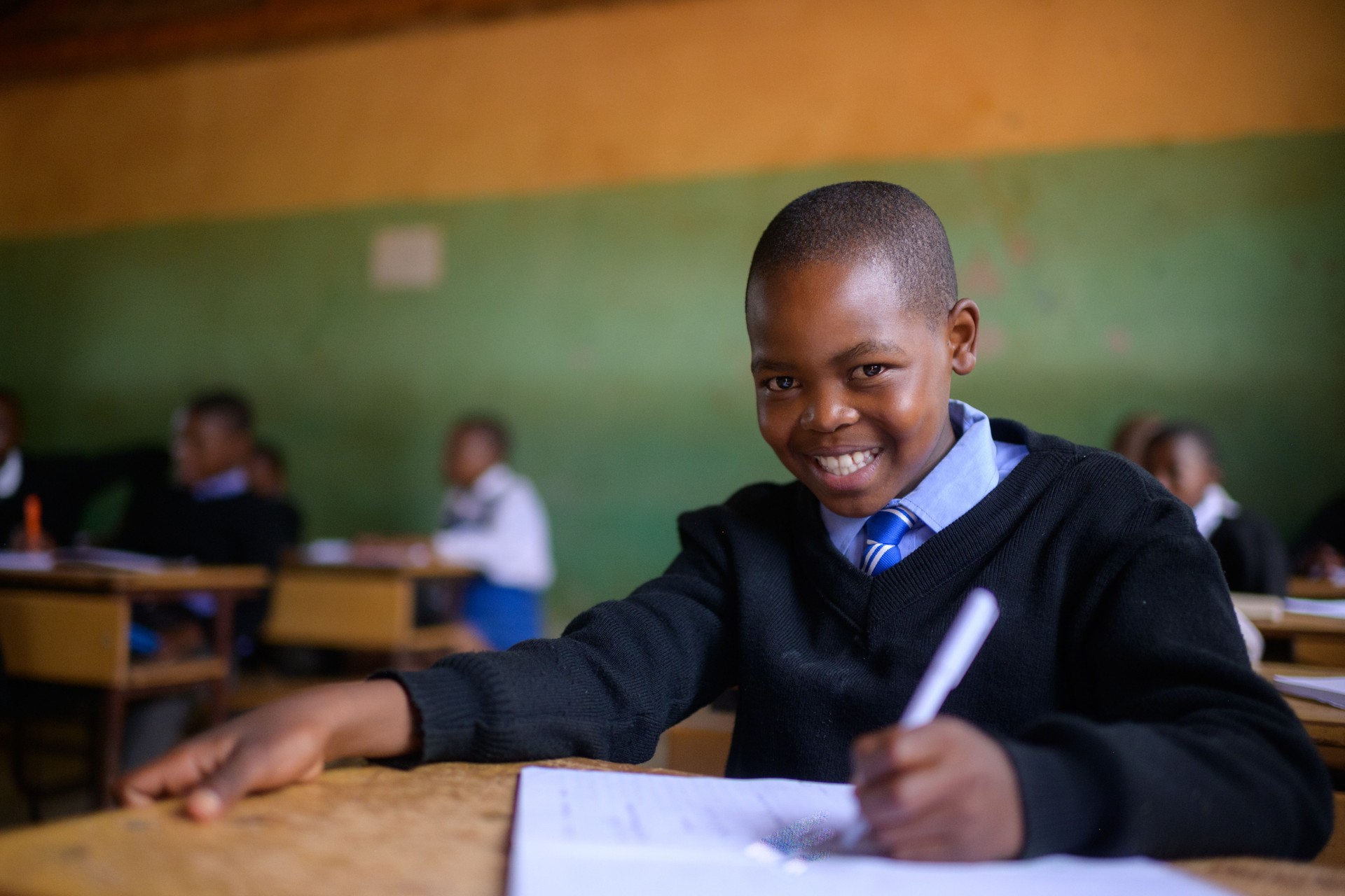 Cute Schoolboy in school uniform sitting in classroom looking at camera smiling and writing